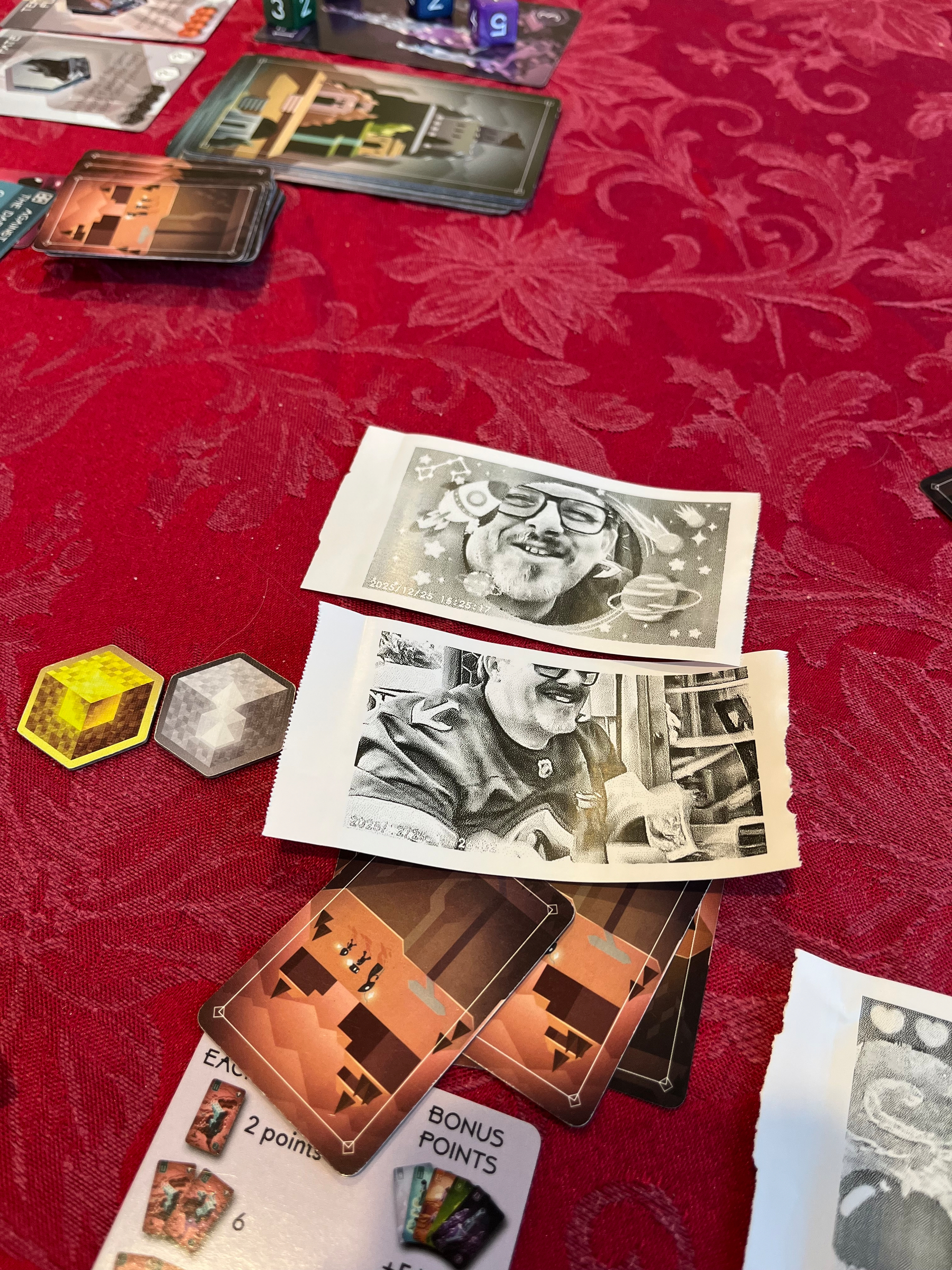 Cards, dice, and stones from the game of Unearth on a table with a couple of black and white pictures of me printed on thermal recipt paper. 